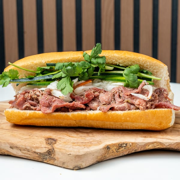 A Banh Mi Ong Beo sandwich with beef, fresh cilantro, and pickled vegetables on a wooden board.