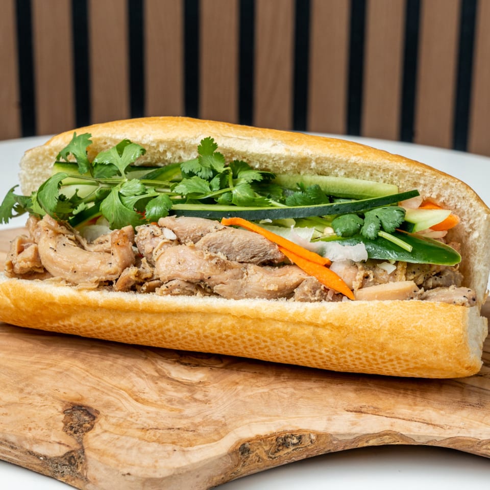 A Banh Mi Ong Beo sandwich filled with meat, cilantro, cucumbers, and pickled carrots on a wooden board.