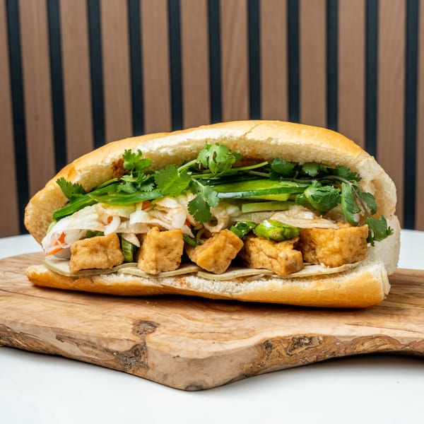 A Banh Mi Ong Beo sandwich with crispy tofu, fresh cilantro, and pickled vegetables on a wooden board.