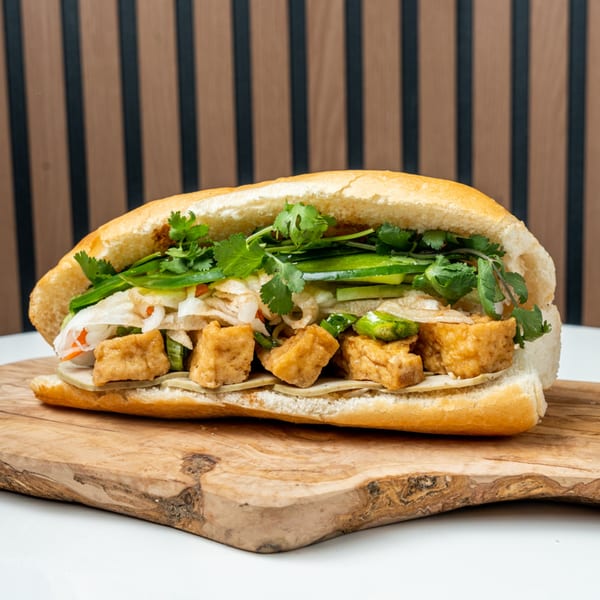 A Banh Mi Ong Beo sandwich with crispy tofu, fresh cilantro, and pickled vegetables on a wooden board.