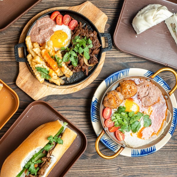 Two sizzling platters with eggs, meat, fries, and tomatoes, plus a banh mi sandwich.