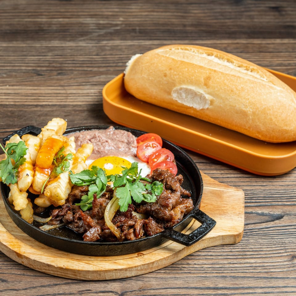 Sizzling beef, fried egg, fries, pate, and tomatoes in a cast iron skillet with a baguette.