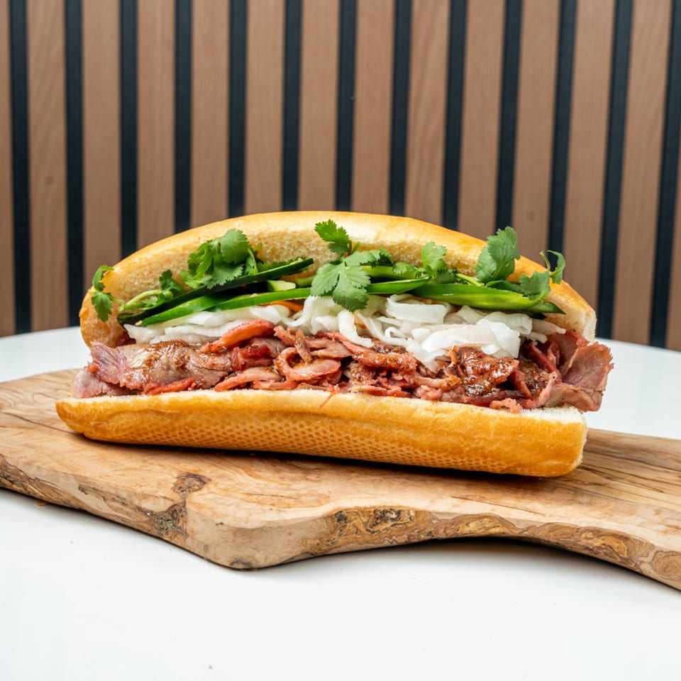 A Banh Mi Ong Beo sandwich with sliced meat, cilantro, and vegetables on a wooden board.