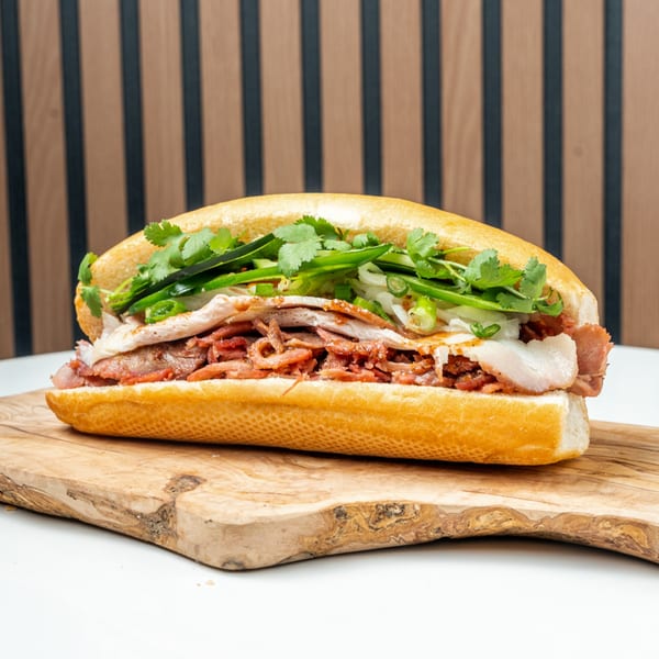 A Banh Mi Ong Beo sandwich, filled with various meats, fresh cilantro, and green onions, on a wooden board.