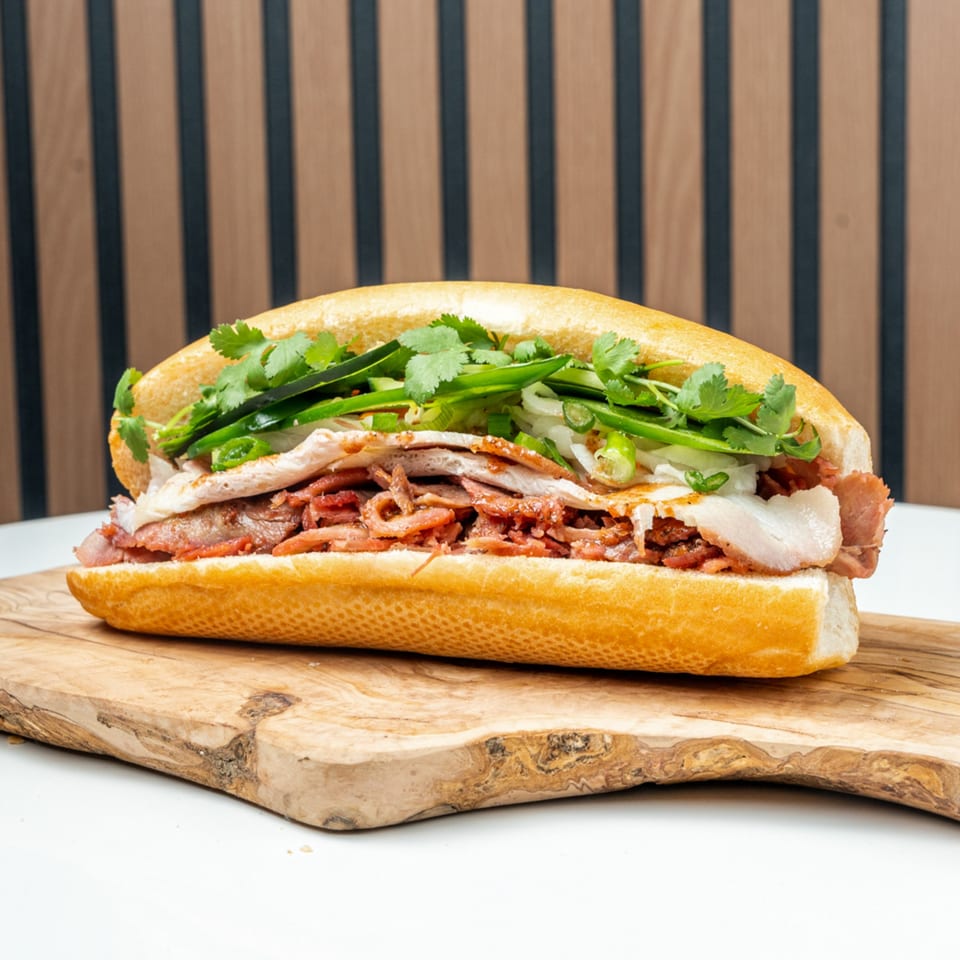 A Banh Mi Ong Beo sandwich, filled with various meats, fresh cilantro, and green onions, on a wooden board.