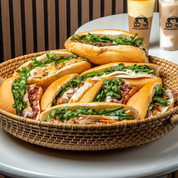 A woven basket filled with various Banh Mi Ong Beo sandwiches and two branded drinks in the background.