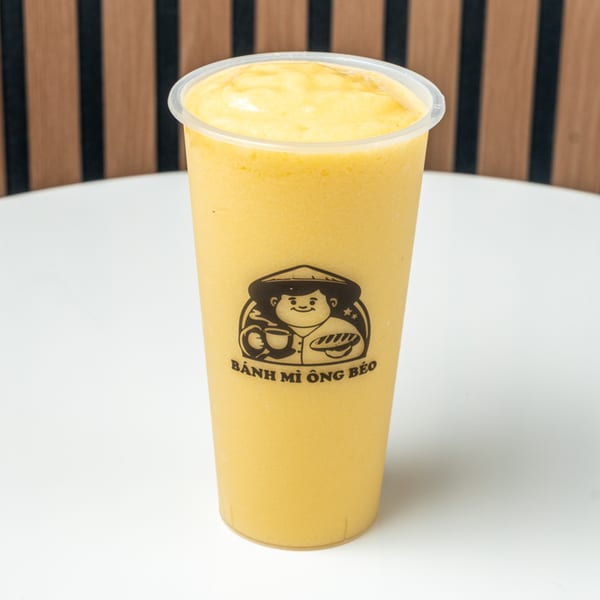 A Banh Mi Ong Beo branded cup filled with a yellow mango smoothie, on a white table.