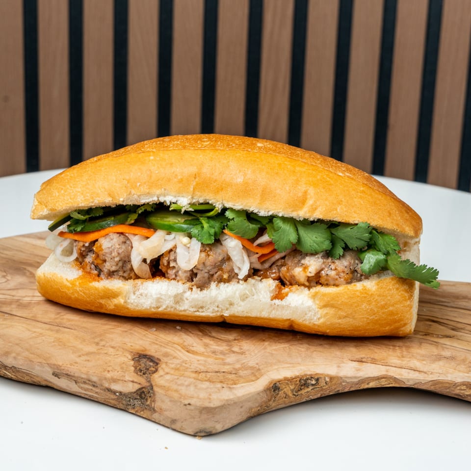 A Banh Mi Ong Beo sandwich with meatballs, pickled vegetables, and cilantro on a wooden board.