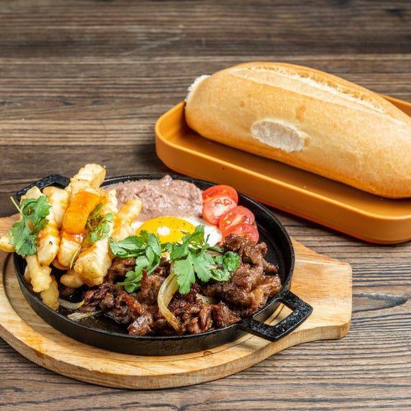 Sizzling beef, fries, fried egg, pate, and tomatoes in a skillet with a banh mi baguette.