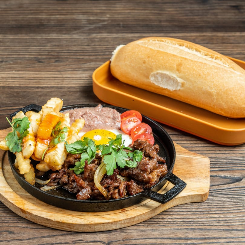 Sizzling beef, fries, fried egg, pate, and tomatoes in a skillet with a banh mi baguette.